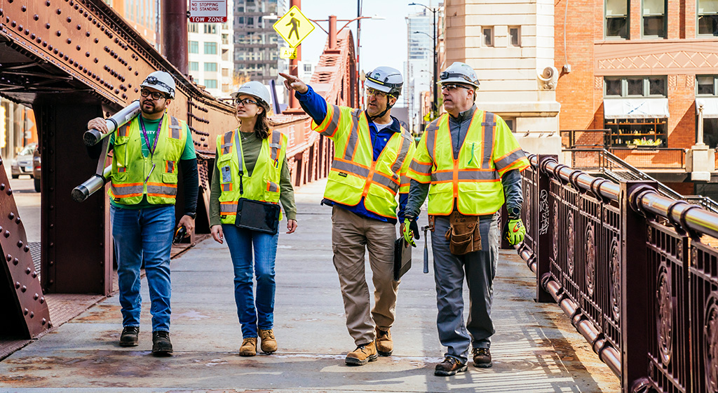 four people in construction hats and vests walking on a bridge and pointing at a building